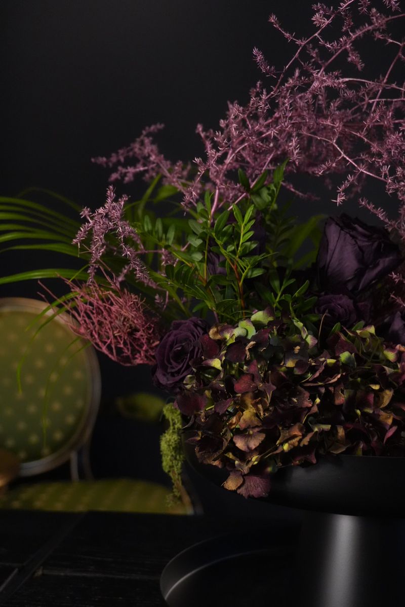 Nahaufnahme eines Arrangements mit Hortensie in Bordeaux und Grün, dunkelvioletten Rosen und rosa gefärbten, feinen Zweigen