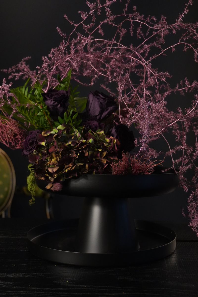Arrangement aus dunkelvioletten Rosen, Hortensie in Bordeaux und Grün und rosa gefärbten, feinen Zweigen auf einer schwarzen Schale