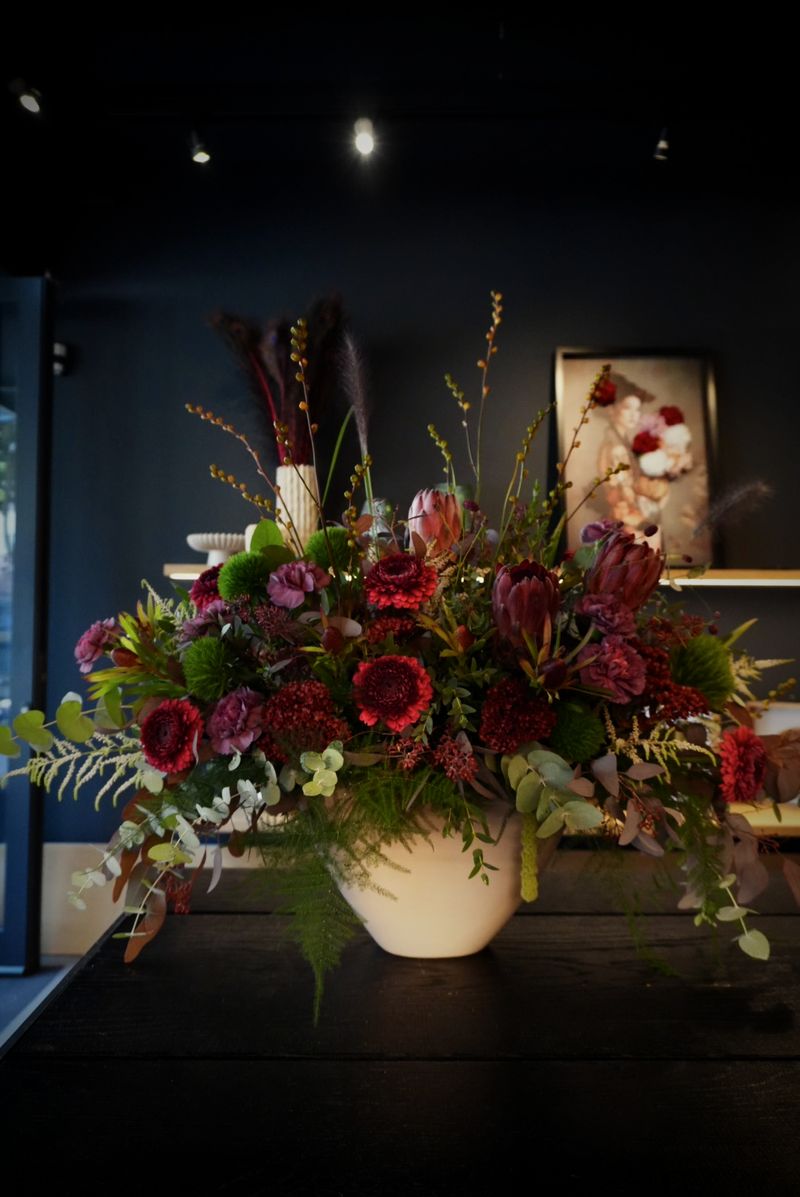 Breites Arrangement mit roten Dahlien, Protea, Eukalyptus und grünen Chrysanthemen in einer weißen Vase
