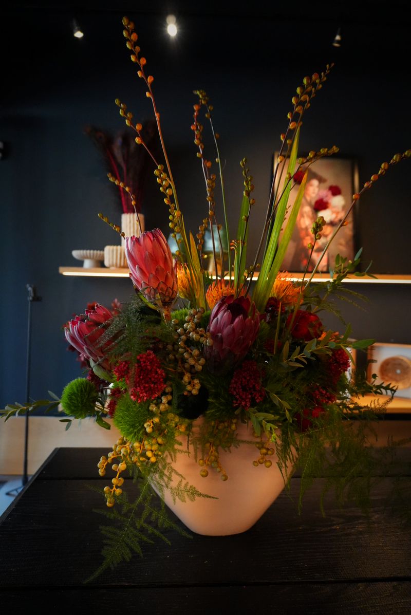 Arrangement mit pinker Protea, roten Blüten und Beerenzweigen in einer hellen Vase