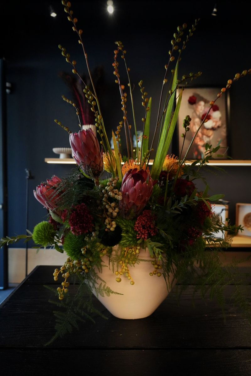 Hohes Arrangement mit pinker Protea, orangefarbenen Blüten, roten Nelken, gelben Beerenzweigen und Gräsern in einer hellen Vase vor den Regalen der Blumengalerie