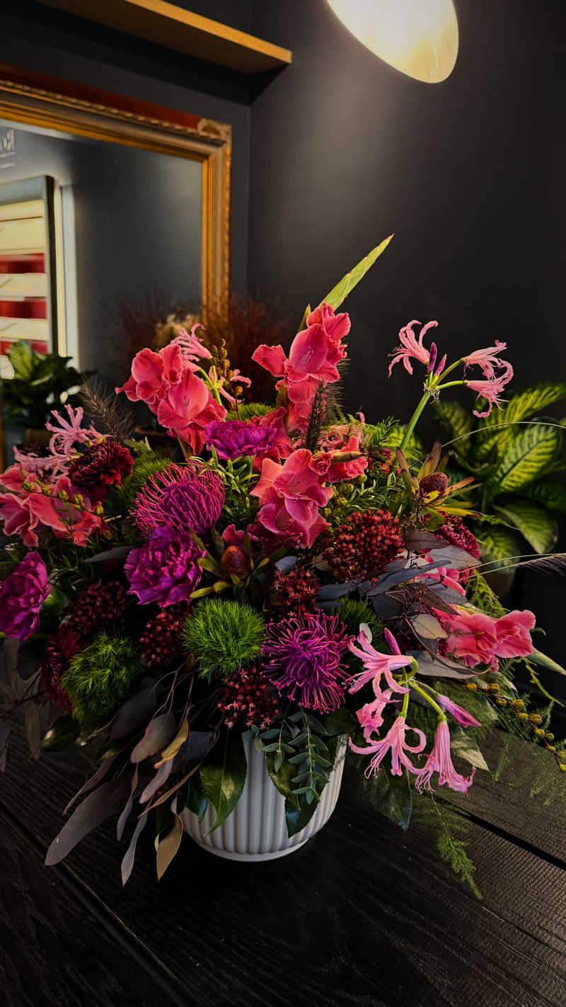 Leuchtend pinkes Tischgesteck mit Nerinen, Gloriosa und Disteln vor einem goldenen Spiegel