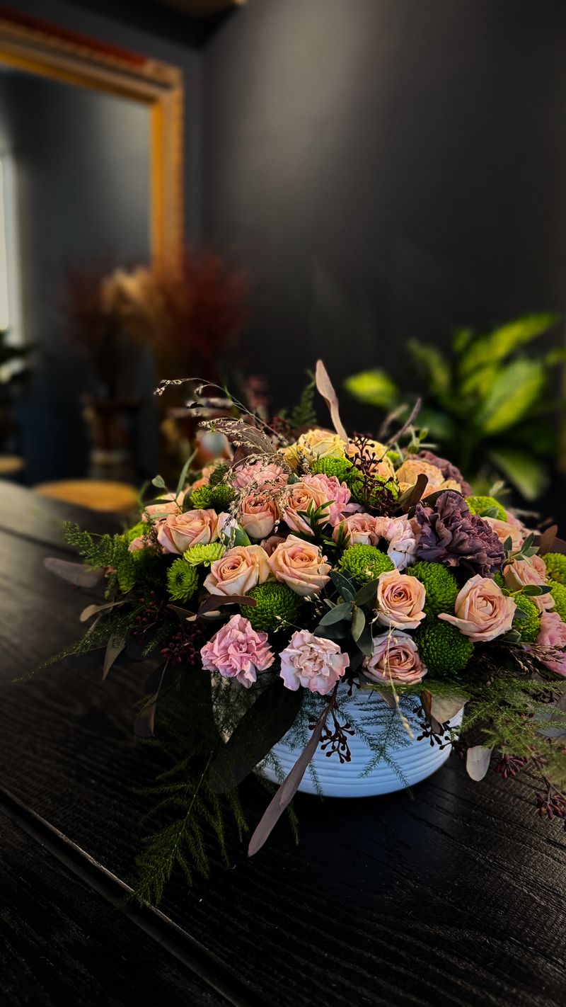 Rundes Gesteck aus pfirsichfarbenen Rosen, rosa Nelken und hellgrünen Chrysanthemen auf einem dunklen Holztisch