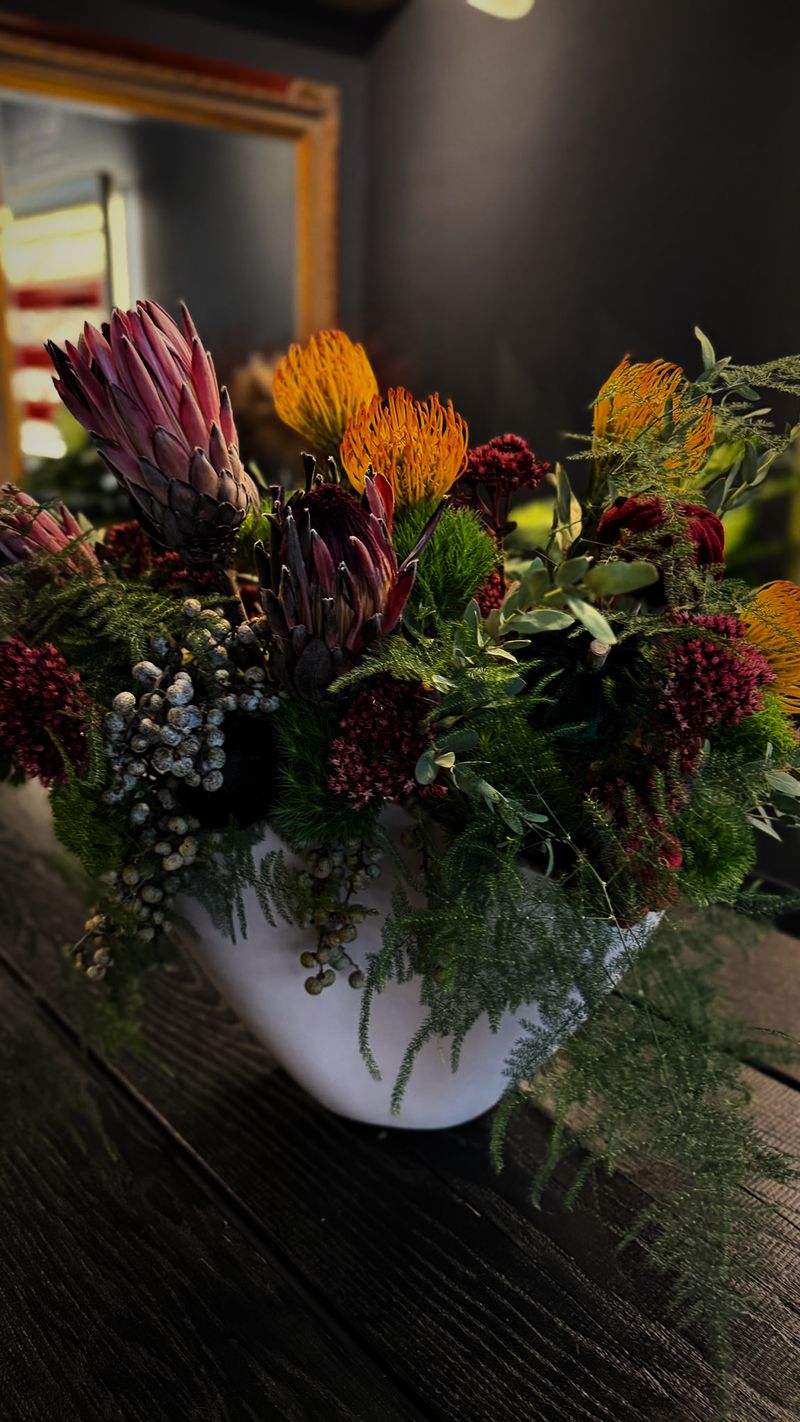 Farbenfrohes Gesteck mit Protea, orangefarbenem Nadelkissen, roten Blüten und Farn in einer weißen Schale